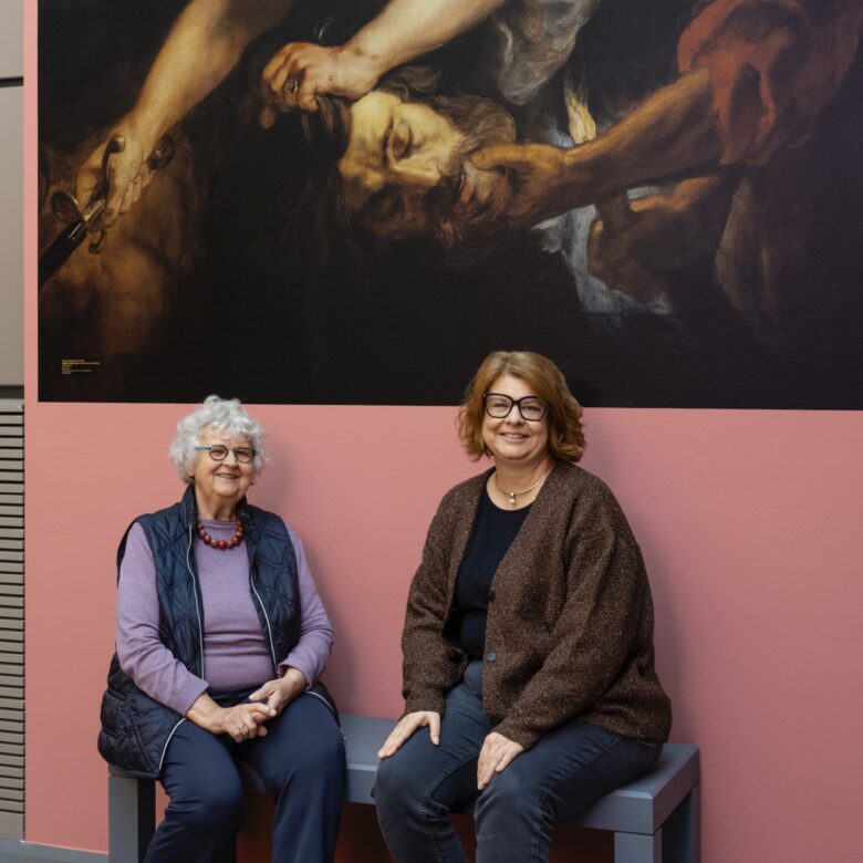 Das Foto zeigt zwei Frauen auf einer Bank im Museum. Im Hintergrund die Reproduktion eines Gemäldes, auf dem zwei Frauen einen Mann köpfen.