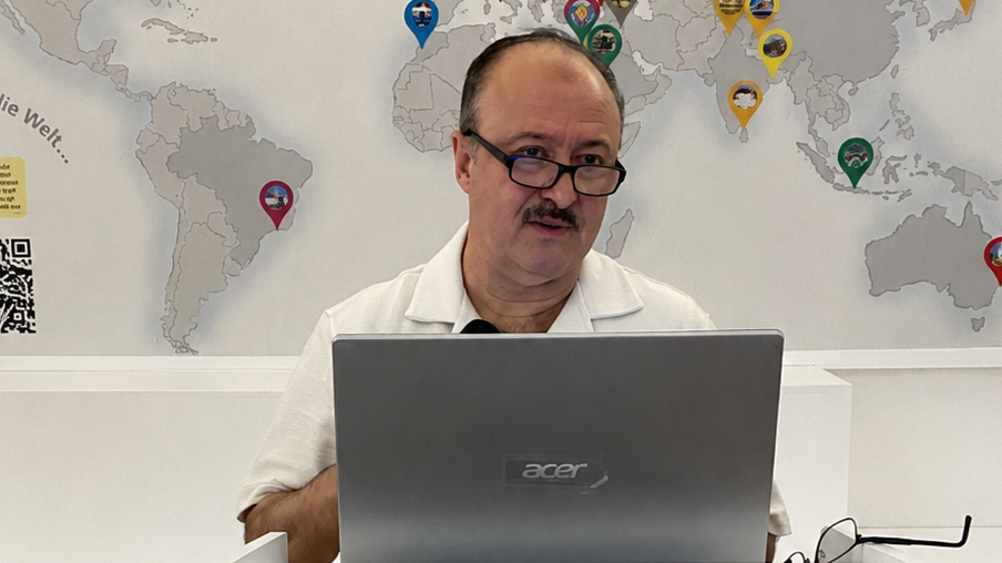 Ein Mann mit Schnurrbart, Brille und zurückweichendem Haaransatz spricht hinter einem Laptop. Im Hintergrund eine Weltkarte.