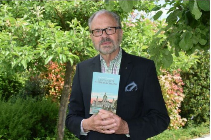 Ein Mann mit grauem Bart und Brille hält ein Buch vor der Brust.