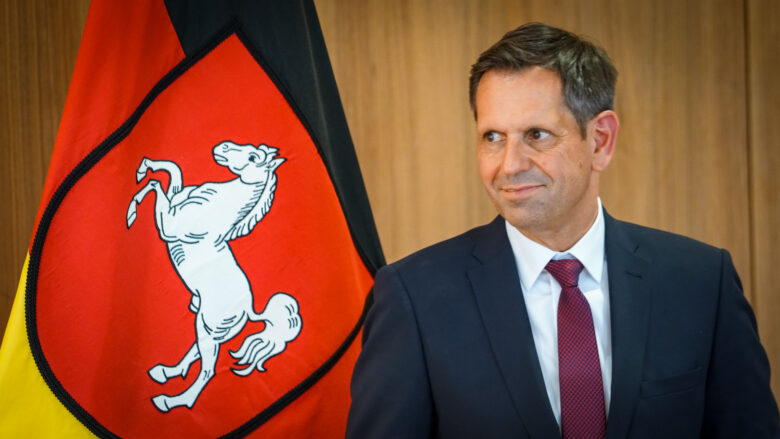 Olaf Lies steht vor der Niedersachsen-Flagge und lächelt verschmitzt. | Foto: Link