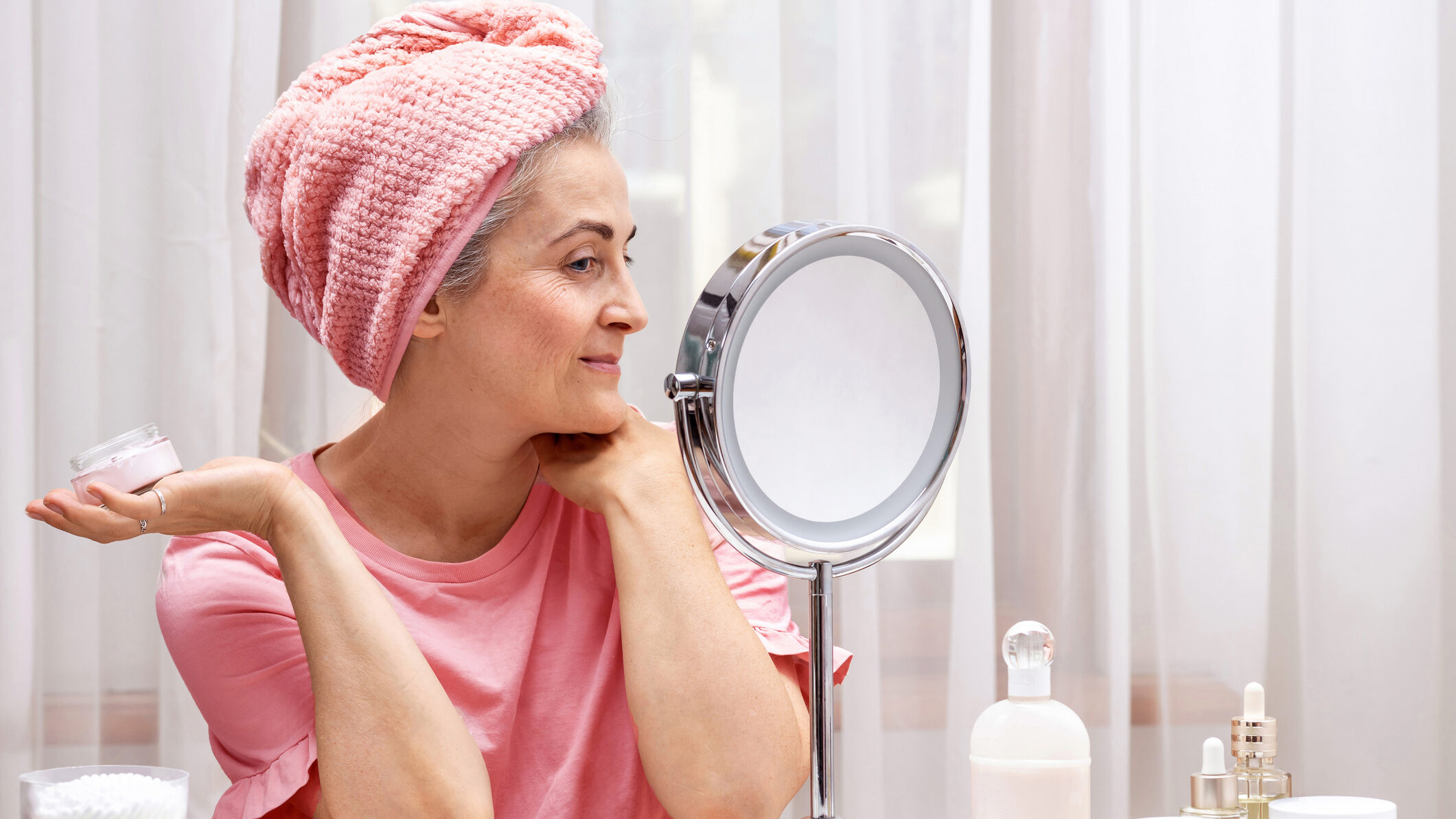 A 50 year old woman in front of a mirror takes care of her face and neck skin while sitting at her dressing table