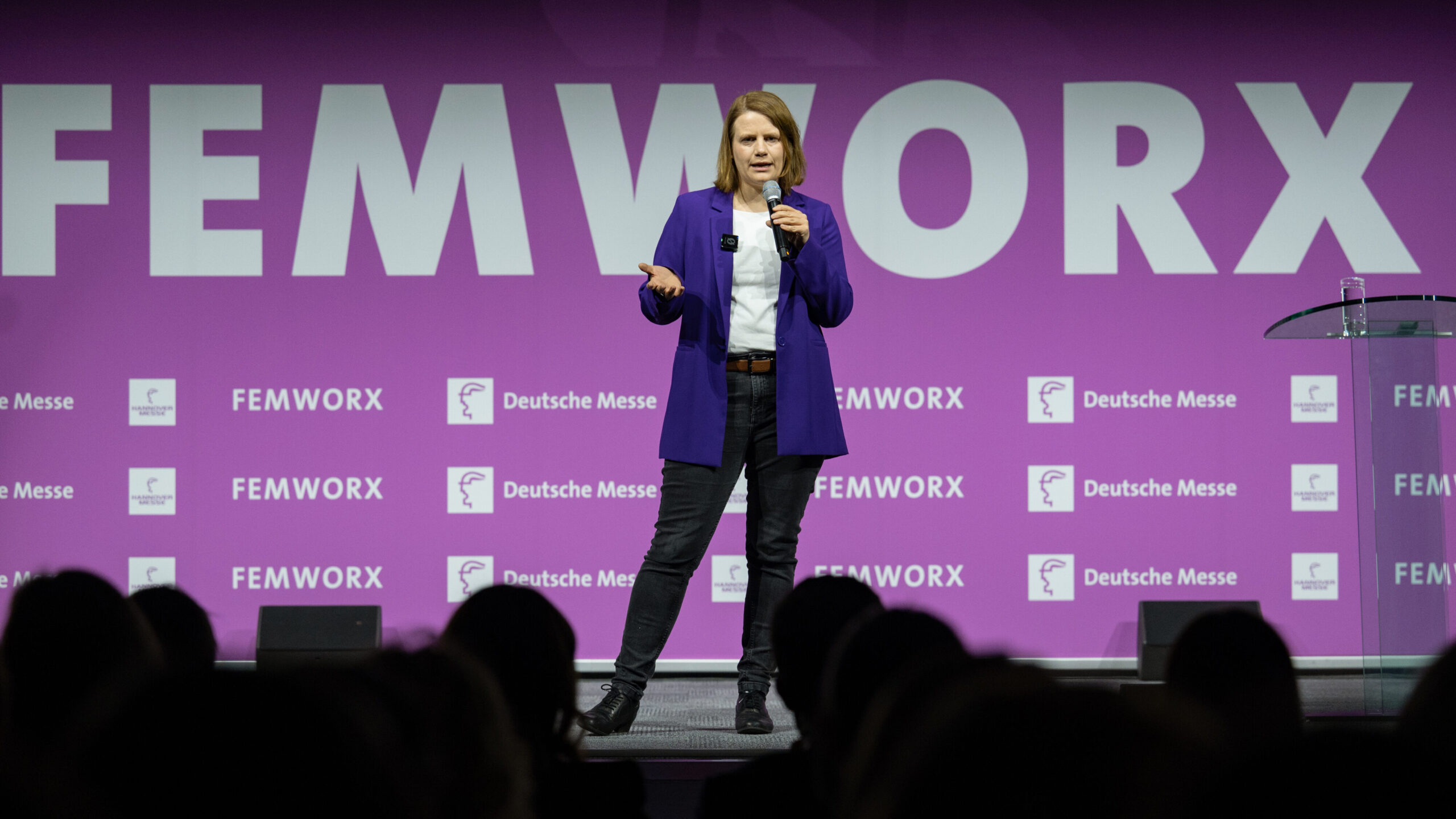 Julia Willie Hamburg steht gestikulierend vor einem pinken Hintergrund mit der Aufschrift "Femworks" und hält ein Mikrofon in der Hand.