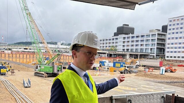 Ein Foto zeigt Minister Falko Mohrs in Warnweste und Bauhelm, im Hintergrund eine große Baugrube und große, funktionale Gebäude aus dem 20. Jahrhundert.