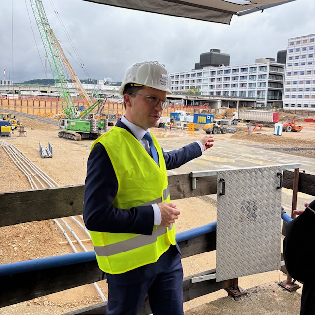 Ein Foto zeigt Minister Falko Mohrs in Warnweste und Bauhelm, im Hintergrund eine große Baugrube und große, funktionale Gebäude aus dem 20. Jahrhundert.