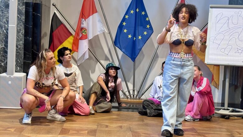Ein Foto zeigt Tänzerinnen in HipHop-Kleidung. Eine steht und spricht, die anderen kauern am Boden.