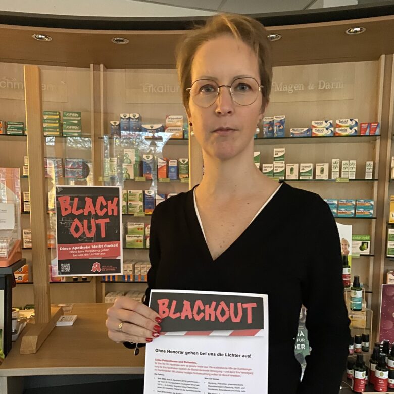 Das Foto zeigt eine Frau mit ernstem Blick in einer Apotheke. Sie hält ein Schild mit der Aufschrift "Blackout".