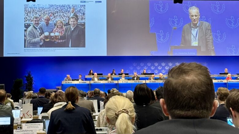 Das Foto zeigt das Plenum des Ärztetages. Die Leinwand ist zweigeteilt. Rechts ist der Redner zu sehen, links Markus Söder, Friedrich Merz, Bärbel Bas und Lars Klingbeil mit Maßkrügen.