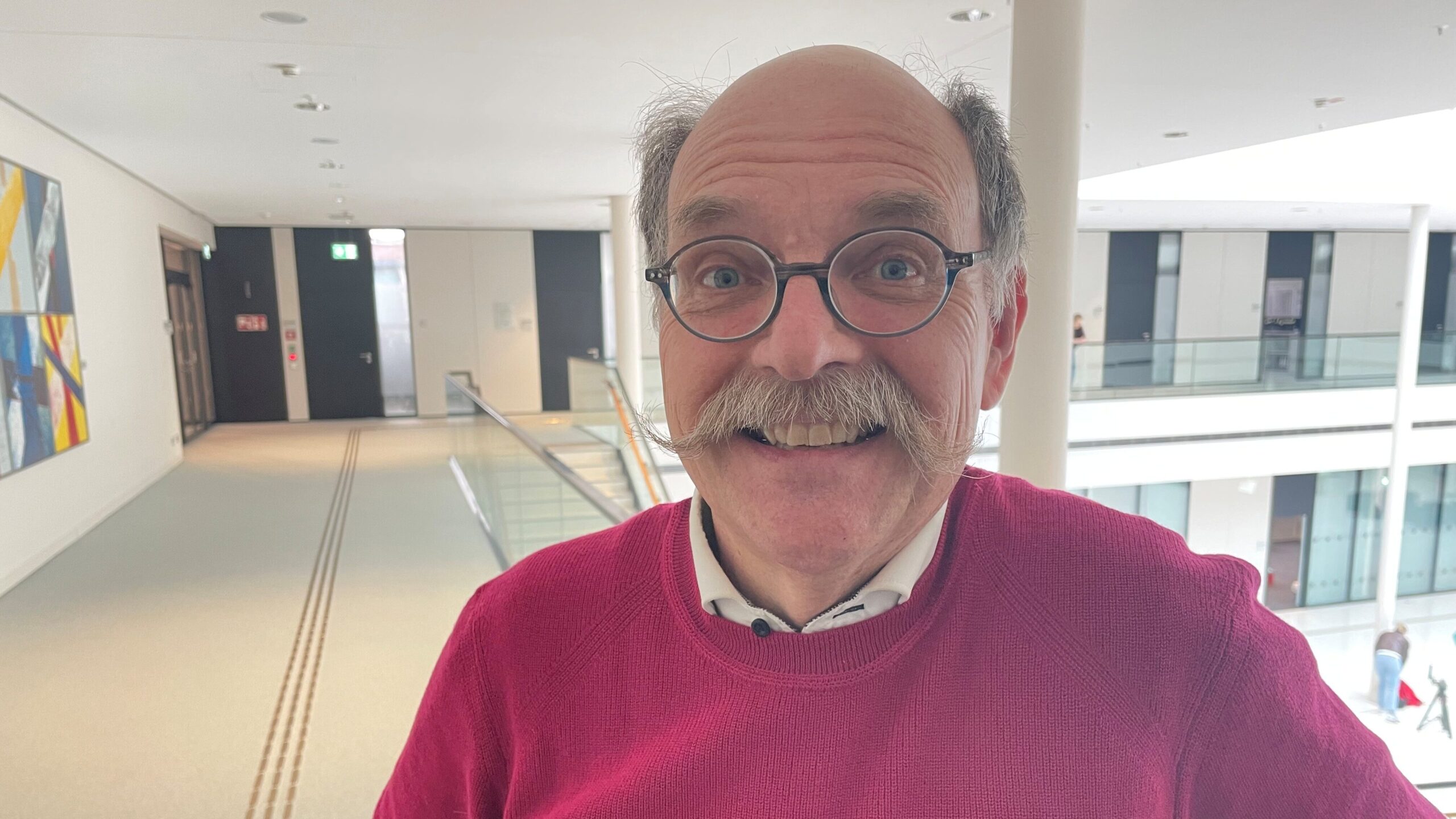 Ein Mann mit grauem Schnurrbart und Glatze reißt die Augen weit auf und lächelt ironisch.