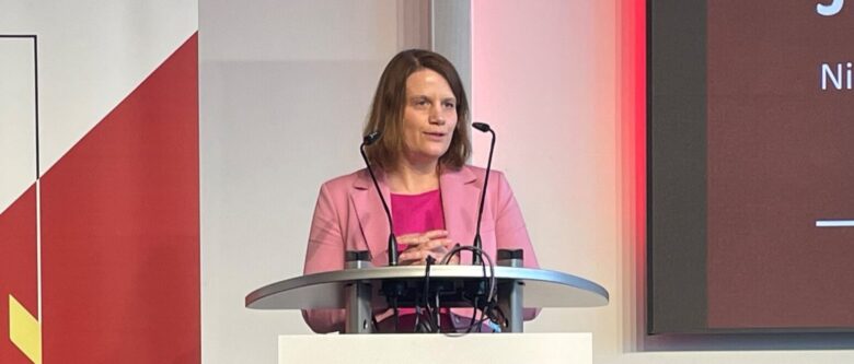 EIne Frau im pinkfarbenen Blazer spricht von einem Rednerpult aus.