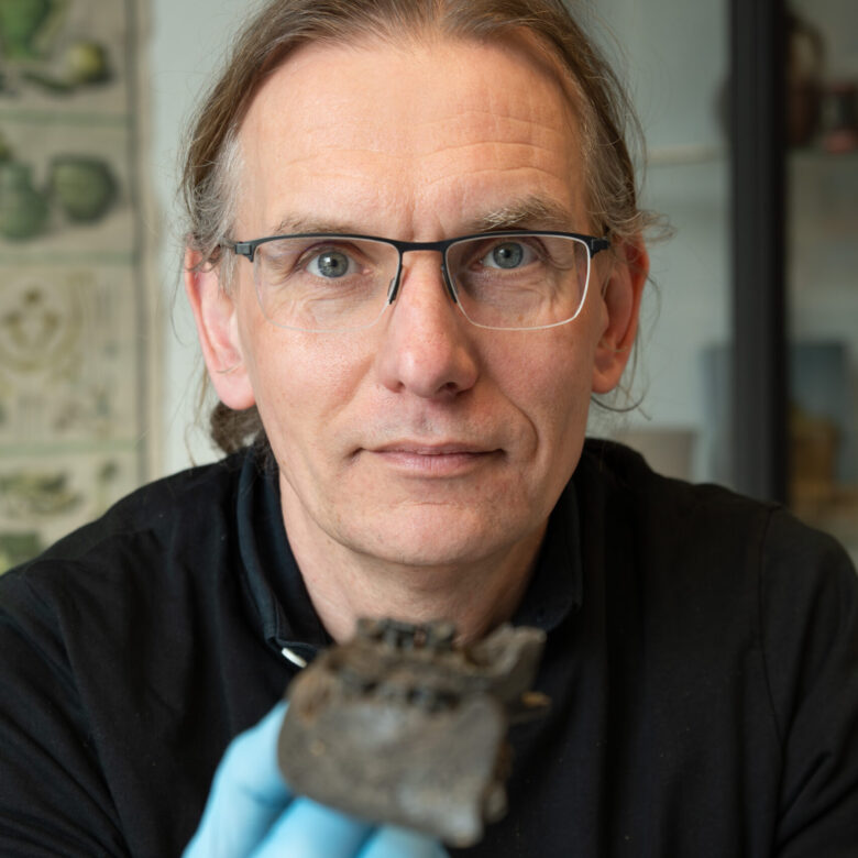 Ein Mann mit Zopf und ernstem Blick hält mit einer behandschuhten Hand ein Knochenstück in die Kamera.
